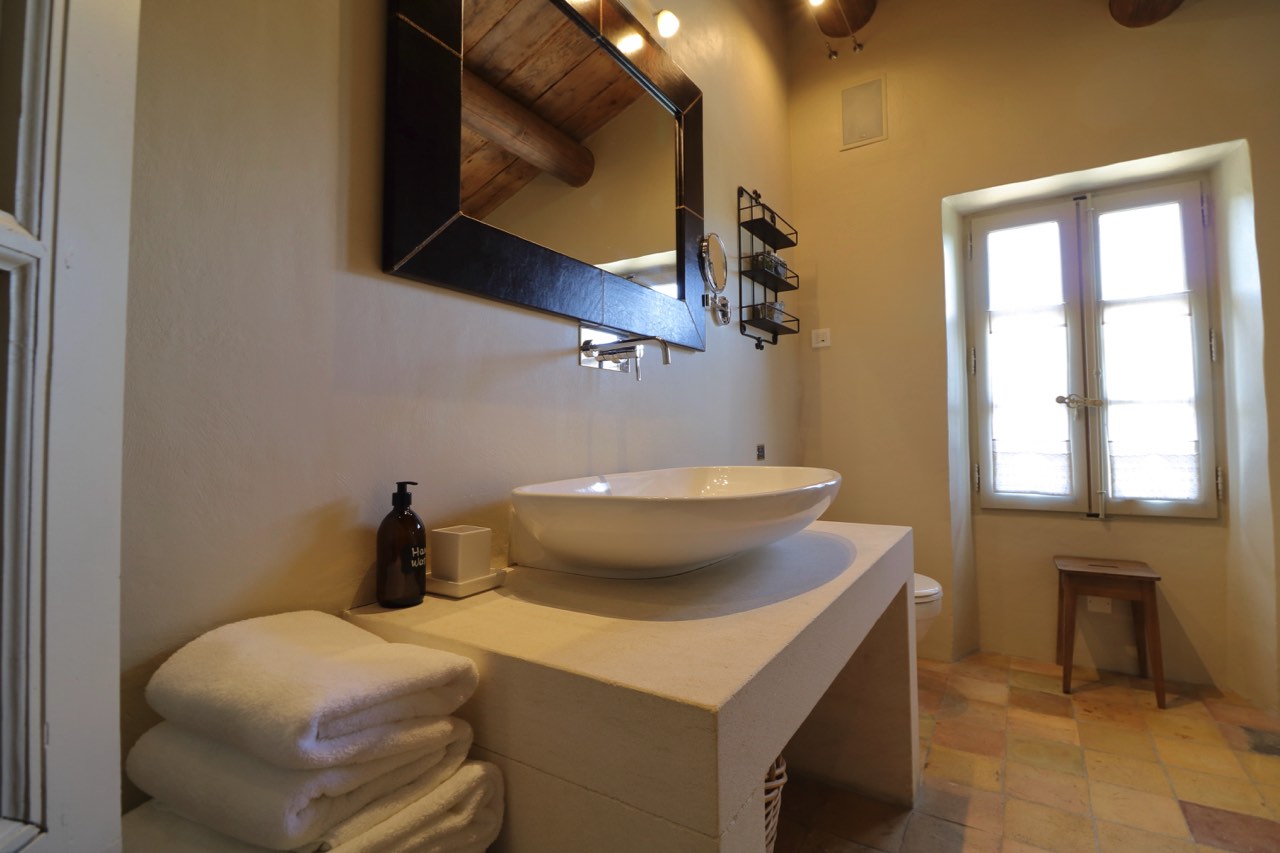 A cozy bathroom featuring a rustic oval sink on a concrete countertop, a large mirror above, and a window with natural light streaming in. Towels are neatly stacked beside the sink.